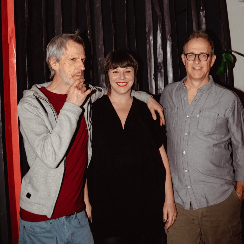 Three people stand in front of a dark, textured background with vertical lines.