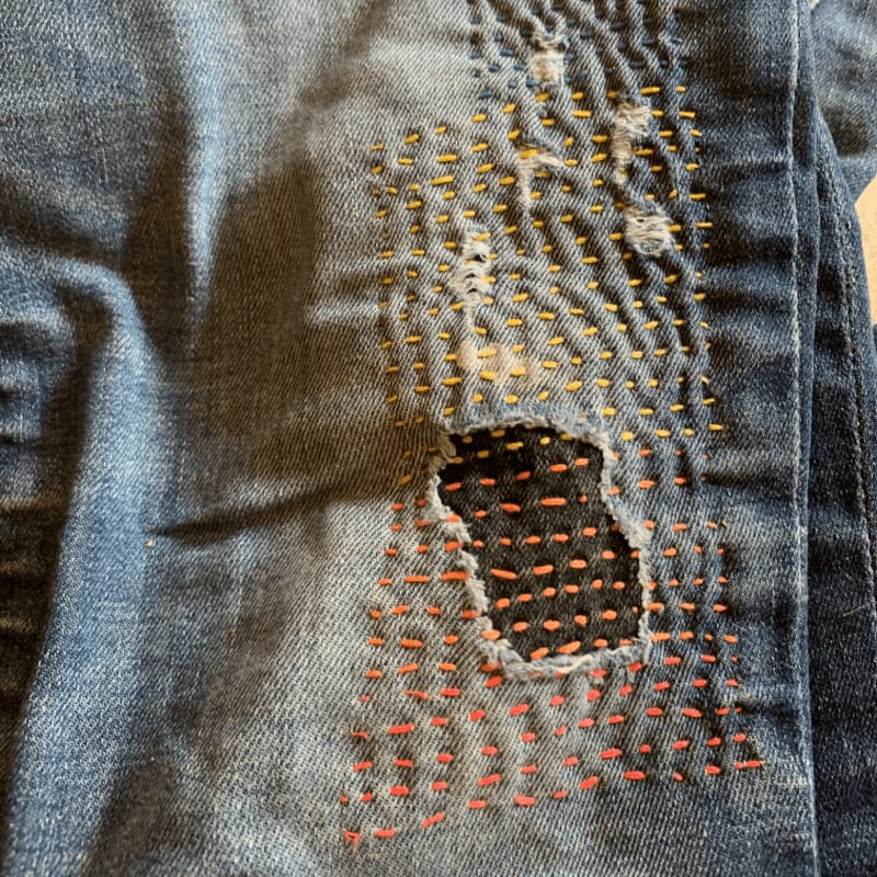 Distressed denim jeans with visible mending stitches in orange and yellow are in the foreground, while the background is out of focus.