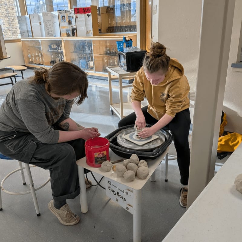 Two young women are working with clay on a pottery wheel in the foreground, while the background shows a room with display cases and trophies.