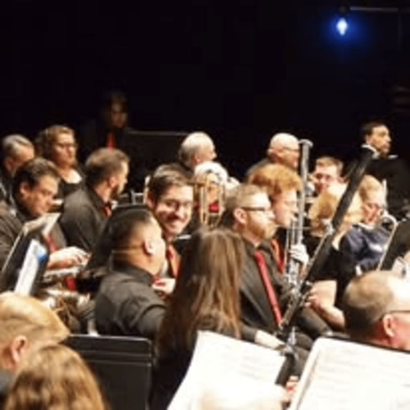 Musicians in formal attire play instruments in a dimly lit concert hall with sheet music visible in the foreground.