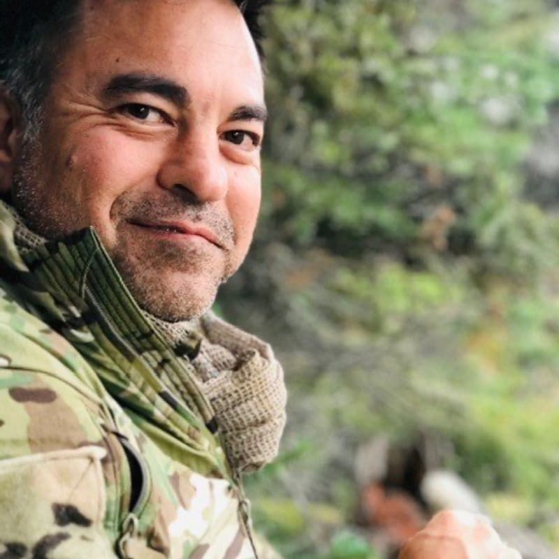 A man in a camouflage jacket smiles at the camera with a blurred forest in the background.