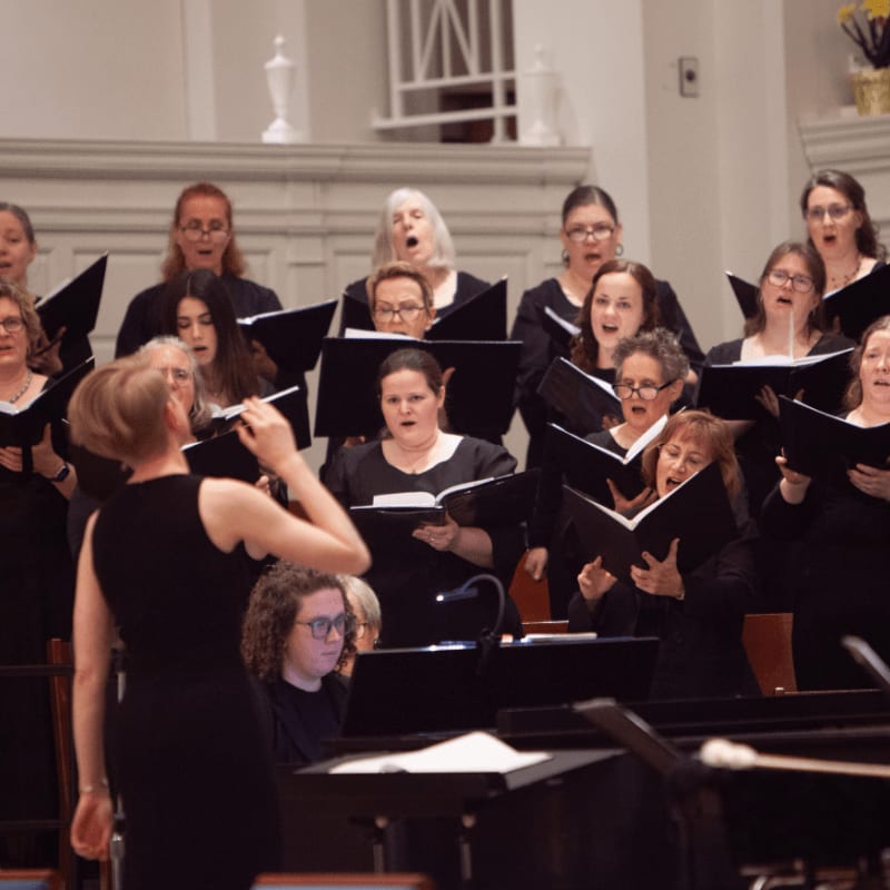 A choir of singers in formal attire performs with sheet music in front of a blurred background of a church interior.