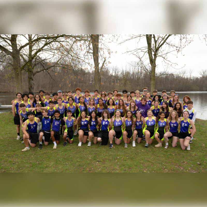 A large group of young athletes in athletic wear poses on a grassy bank in the foreground, with a calm body of water and bare trees in the background.