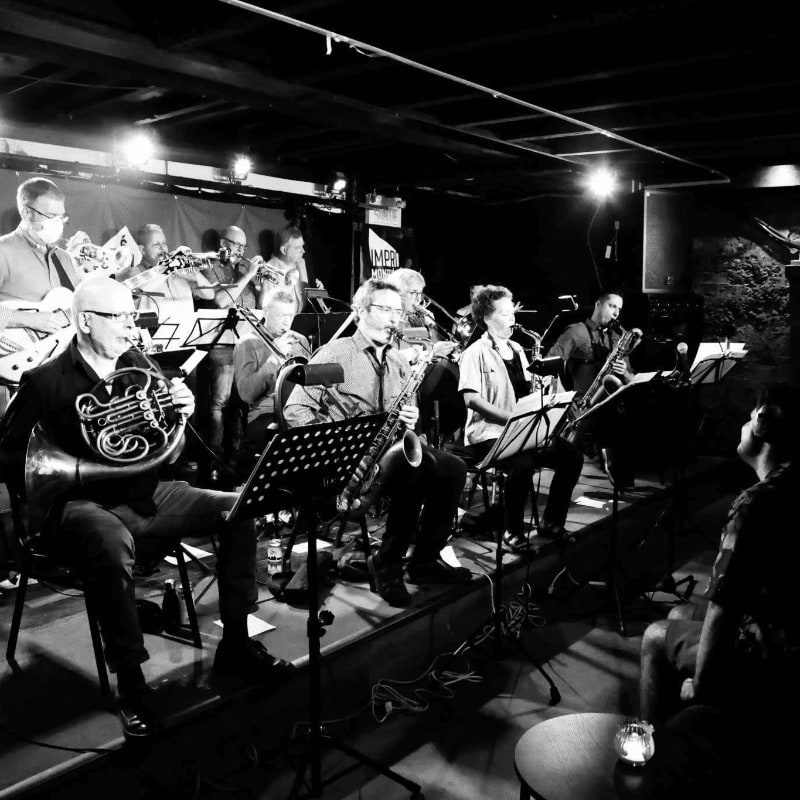 A jazz band performs on a dimly lit stage in the foreground, with audience members visible in the shadowy background.