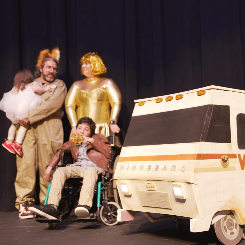 A group of people in costumes, including a child in a wheelchair, stand in front of a cardboard Winnebago prop against a dark stage curtain.