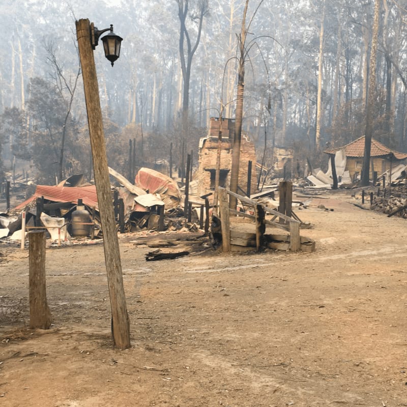 A weathered wooden lamppost stands in the foreground amidst the charred ruins of buildings and trees in the background.