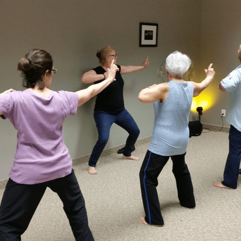 Four people practice Tai Chi in a room with a framed picture on the wall and a spotlight illuminating the background.