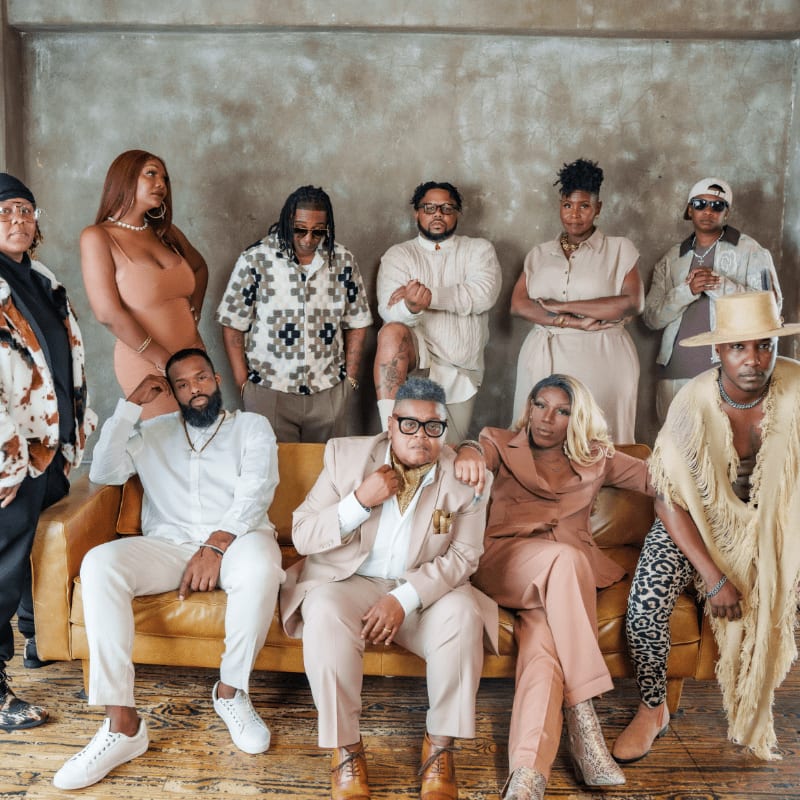 A group of ten stylishly dressed individuals pose in front of a textured gray wall, with some seated on a tan leather couch in the foreground.