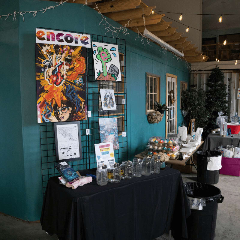 Colorful artwork hangs on a teal wall behind a table displaying jars and books, with a Christmas tree and other vendor tables visible in the background.