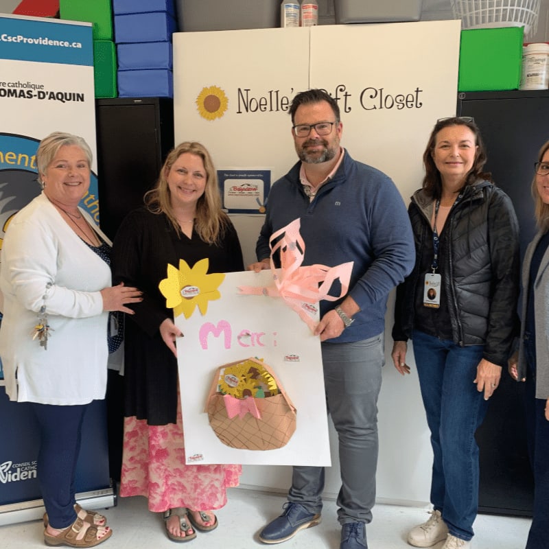 Five people stand in front of a white wall with "Noelle's Craft Closet" written on it, holding a large "Merci" sign decorated with sunflowers and a basket.