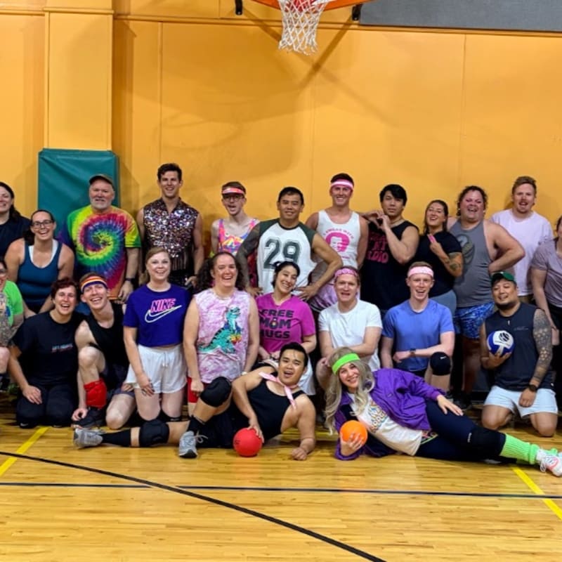 A group of adults in athletic wear poses for a photo in a gymnasium with a basketball hoop visible in the background.