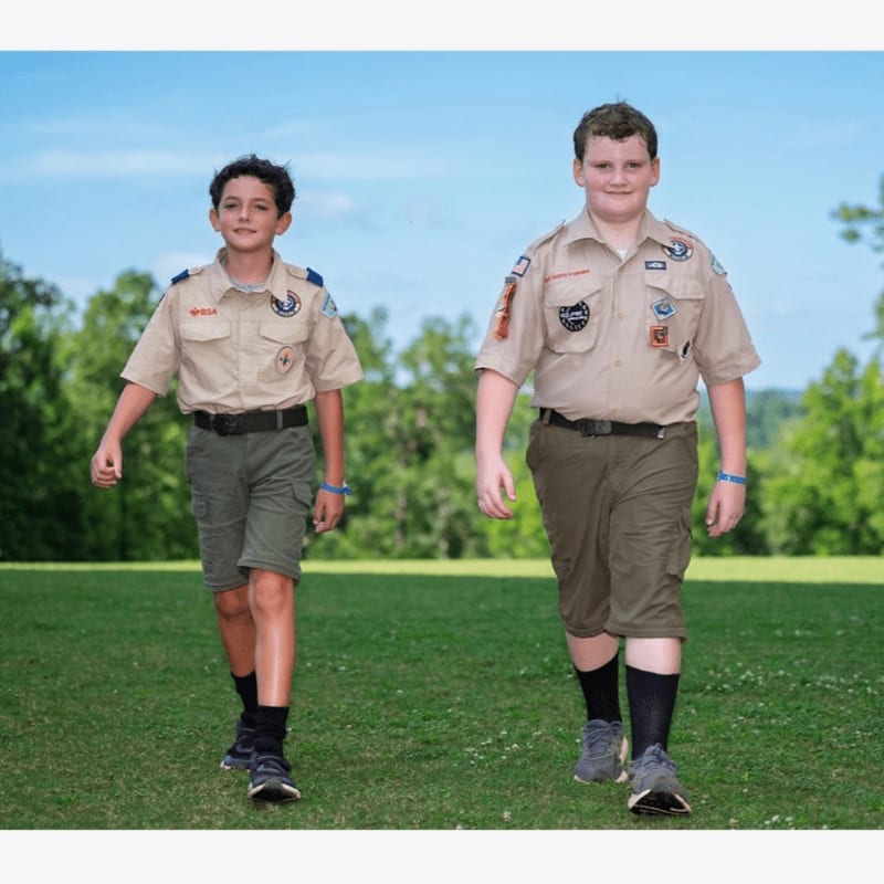 Two young boys in scout uniforms walk across a grassy field with trees and a blue sky in the background.