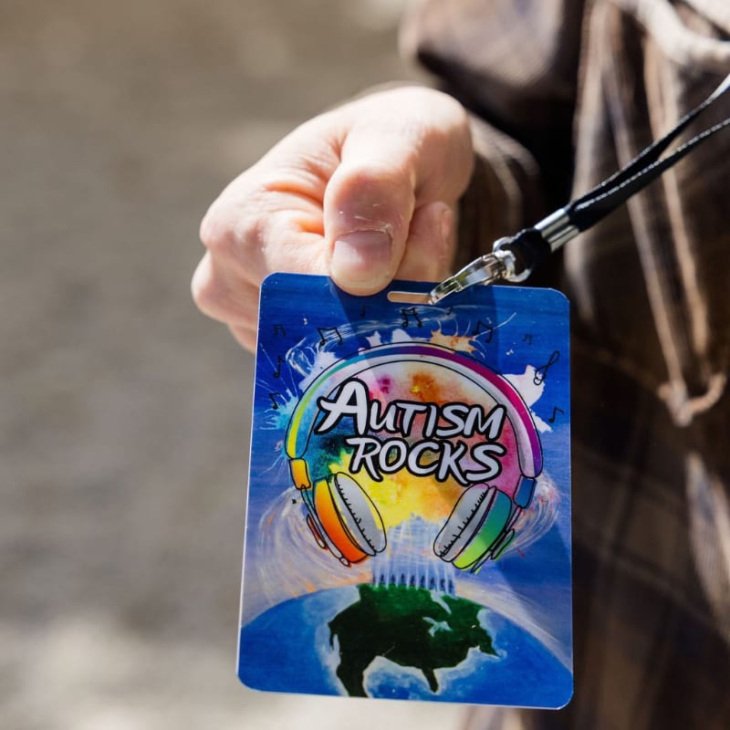 A hand holds up an "Autism Rocks" lanyard with colorful headphones and a globe in the background.