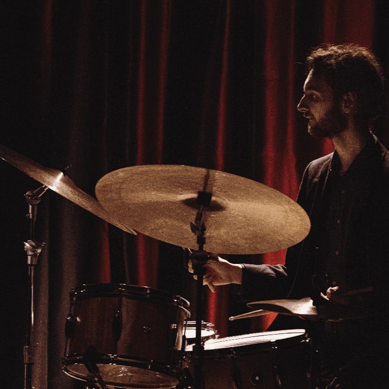 A drummer plays his cymbals and drums in front of a dark red curtain.