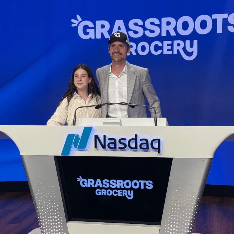 A man and a young girl stand behind a Nasdaq podium with the Grassroots Grocery logo displayed on a blue background.