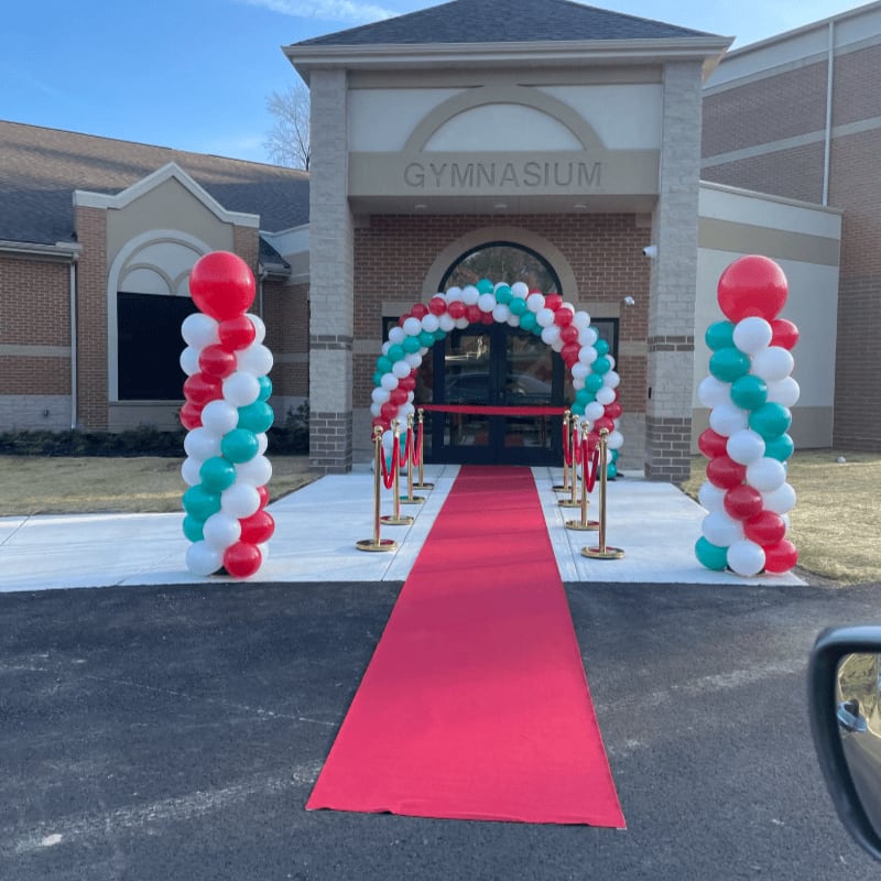 A red carpet leads to a gymnasium entrance adorned with balloon arches and columns, with a brick building and clear sky in the background.