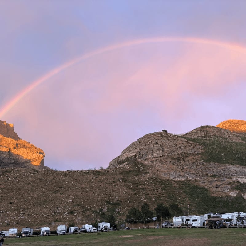A vibrant rainbow arches over a campsite filled with RVs, set against a backdrop of sun-kissed mountains.