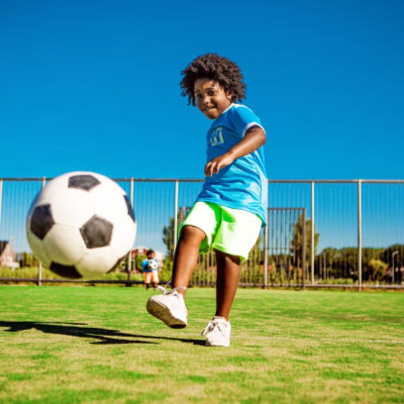 A young boy in a blue shirt and green shorts kicks a large soccer ball on a grassy field with a fence and trees in the background under a clear blue sky.