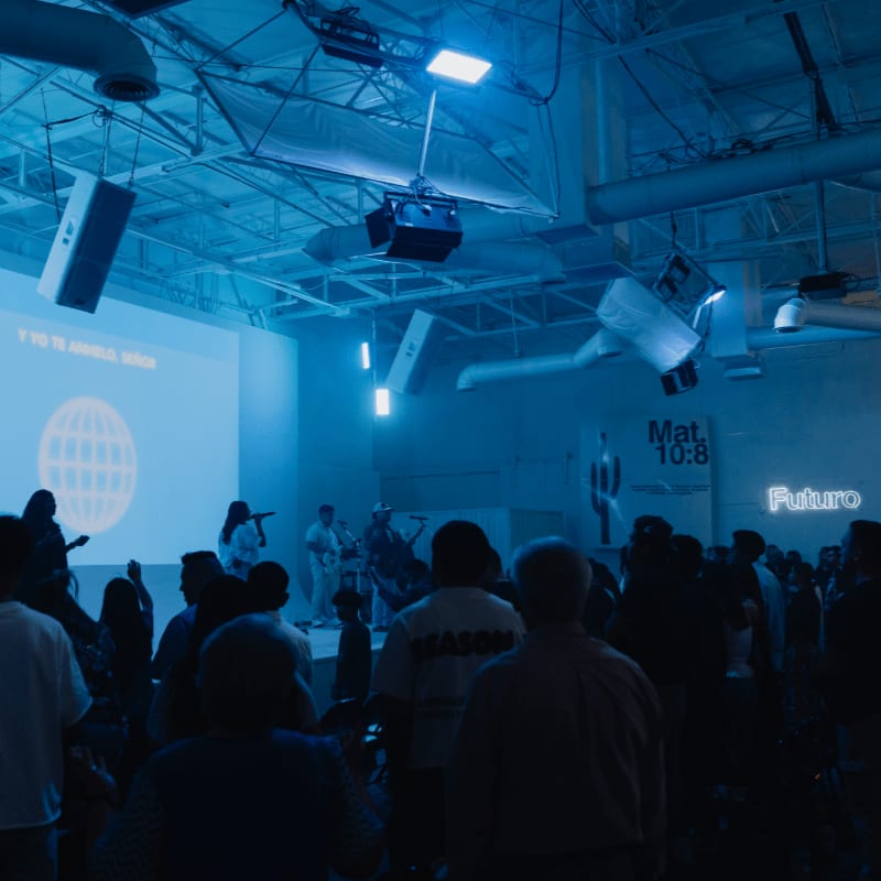 A crowd of people in the foreground watches a band perform on a stage with a projection screen in the background, all bathed in blue light.