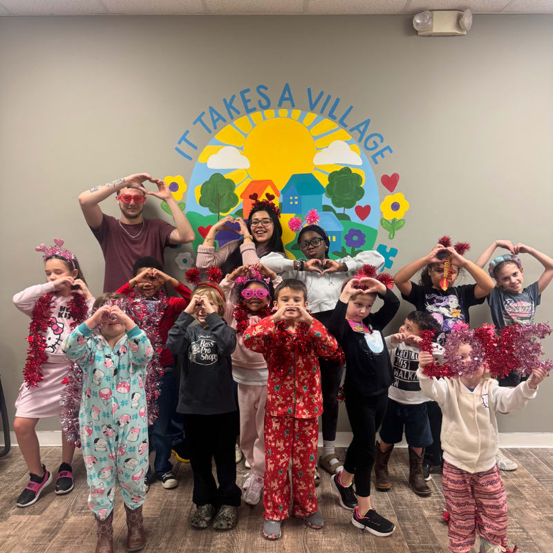 A group of children and adults in pajamas form a heart shape with their hands in front of a mural that reads "It Takes a Village."