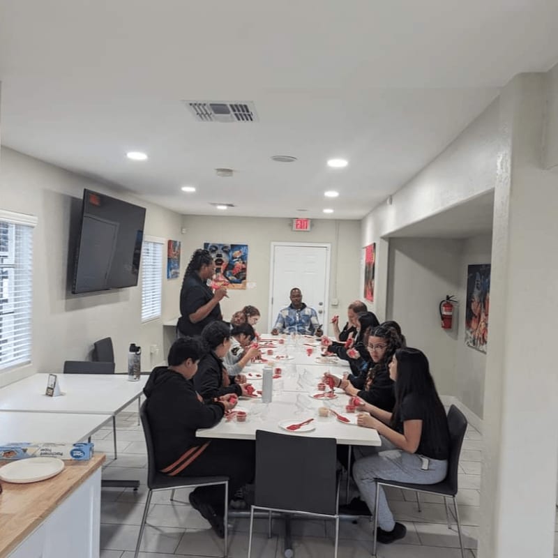 A group of people are gathered around a long table in a brightly lit room, with artwork on the walls and a television mounted on the left.