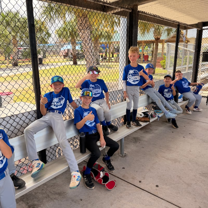 A group of young boys in blue baseball uniforms sit on a bench in the foreground, with a baseball field and palm trees visible in the background.