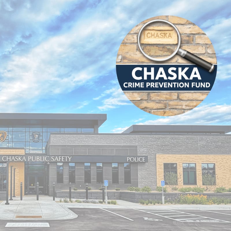 A magnifying glass hovers over the word "CHASKA" on a brick wall, superimposed over a modern public safety building with a cloudy sky in the background.