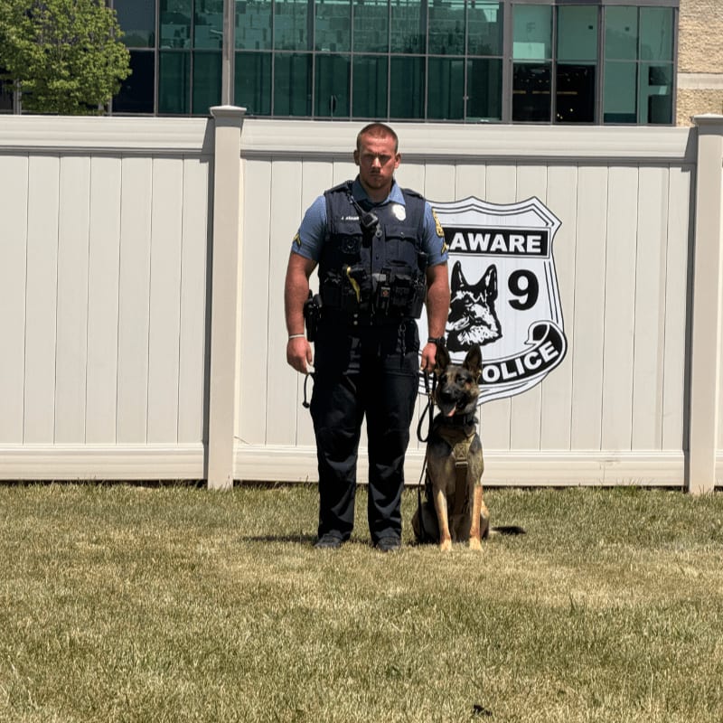 A police officer stands with his K-9 partner in front of a white fence with a police emblem, with a modern building in the background.
