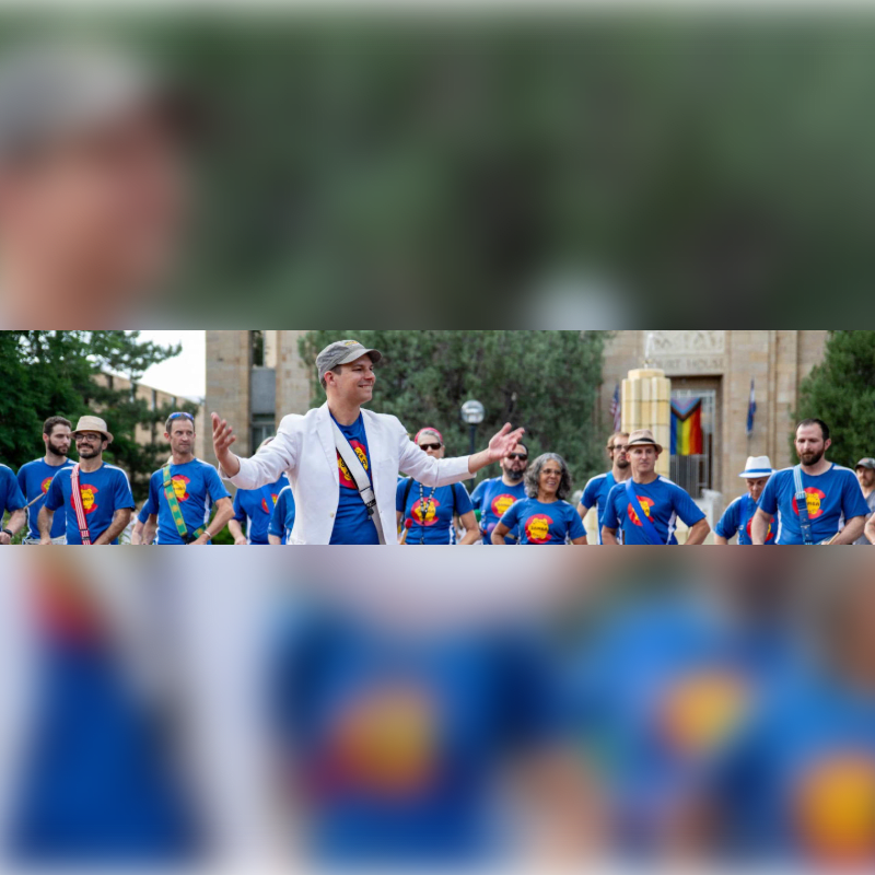 A man in a white jacket leads a group of people in blue shirts with a rainbow emblem, with a building and trees in the background.