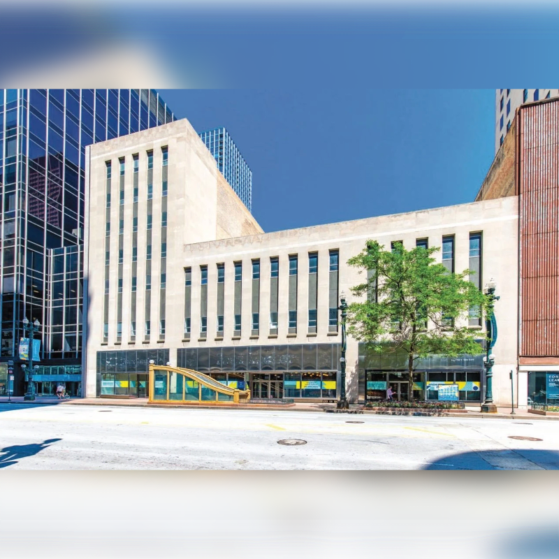 A light-colored, modern building with large windows stands on a bright, empty street, surrounded by taller, glass-fronted skyscrapers.