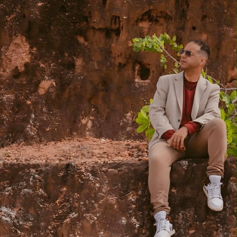 A man in a light-colored suit and sunglasses sits on a rocky ledge with lush green foliage in the background.