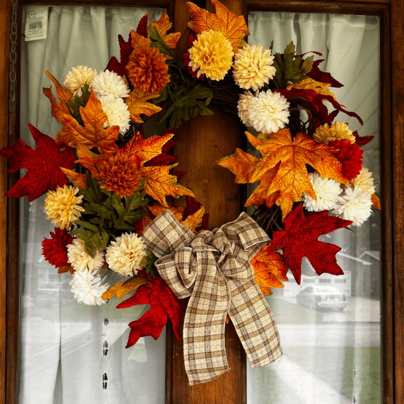A festive fall wreath adorned with colorful leaves and flowers, tied with a plaid bow, hangs on a glass door with sheer curtains in the background.