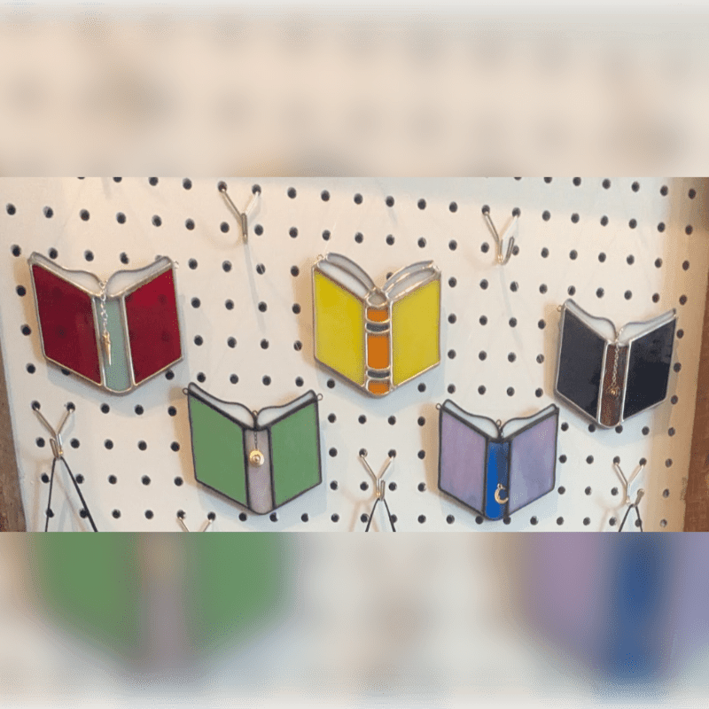 Six colorful stained-glass open books hang from hooks on a white pegboard.