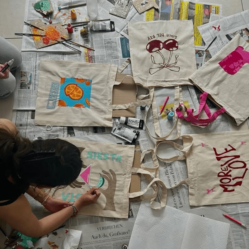 A person is painting a tote bag with a cat design in the foreground, while several other decorated tote bags and art supplies are spread out on newspapers in the background.
