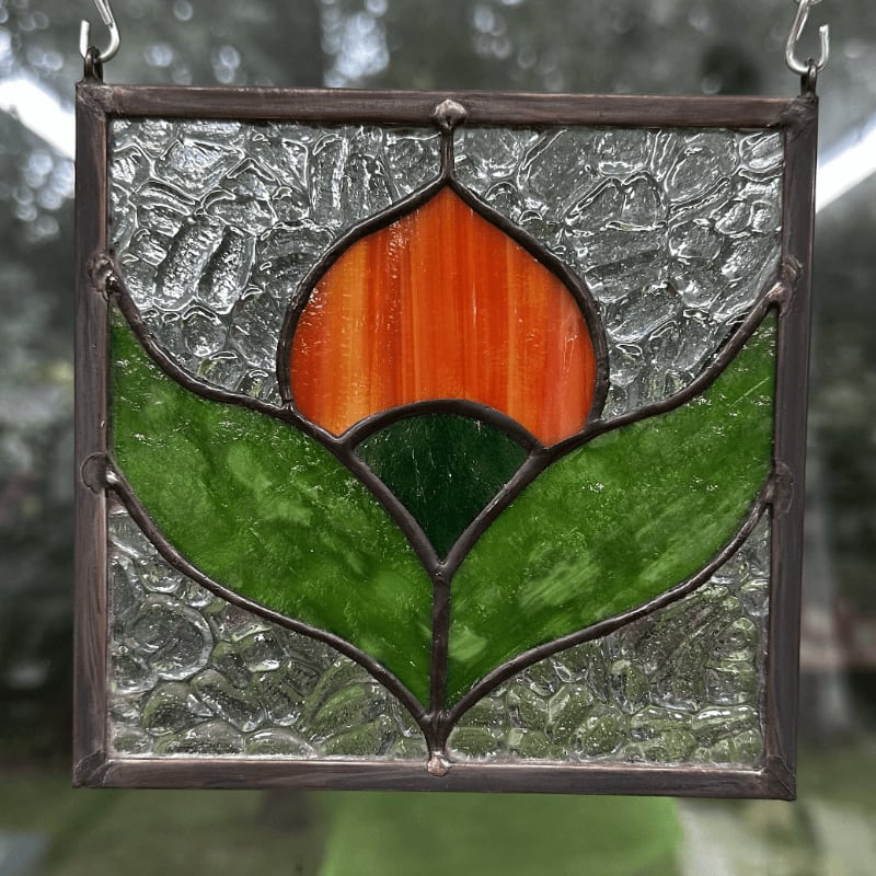 A square stained glass panel featuring an orange and green tulip design hangs in front of a blurred outdoor background.
