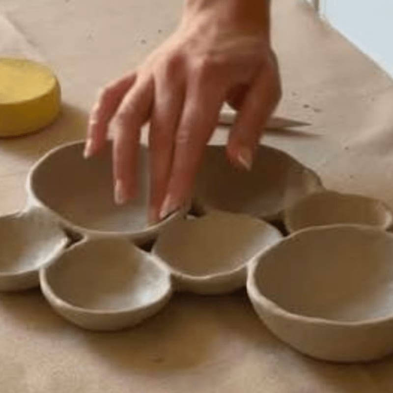 A hand gently touches a cluster of small, unglazed ceramic bowls arranged on a light brown surface, with a yellow tool visible in the blurred background.