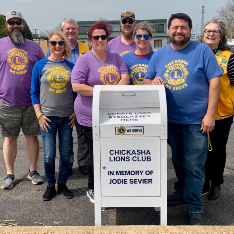A group of people stand behind a donation bin for used eyeglasses, with a building and trees in the background.