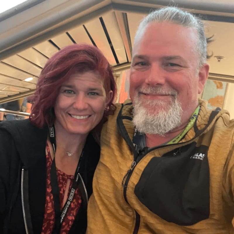 A smiling couple with red hair and a beard pose for a selfie in front of a blurred background with a map.