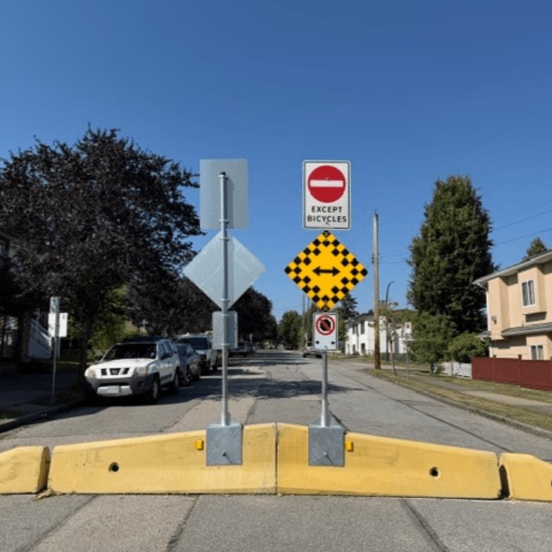 Yellow concrete barriers block a street lined with parked cars and houses under a clear blue sky.