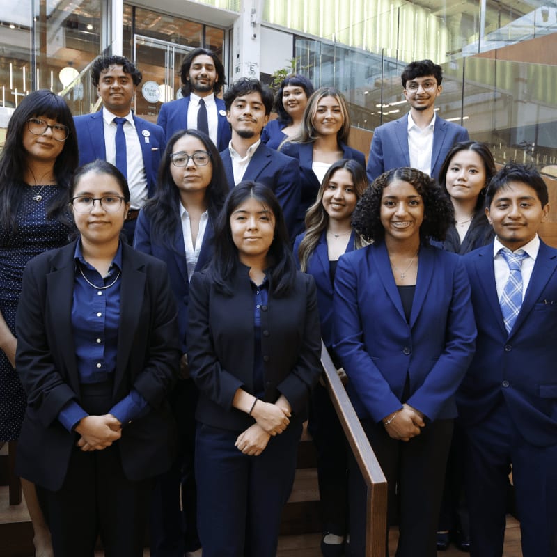 A group of young adults in formal attire stand on stairs in a modern building with large windows in the background.