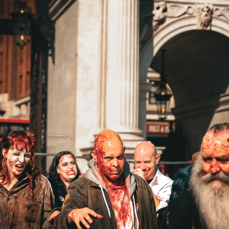 A group of bloody zombies with vacant stares walk towards the viewer in front of an ornate building with arched doorways.