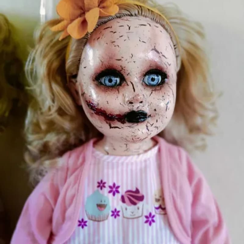 A creepy doll with cracked skin and bright blue eyes wears a pink cardigan over a cupcake-themed shirt, with a blurry, light-colored background.