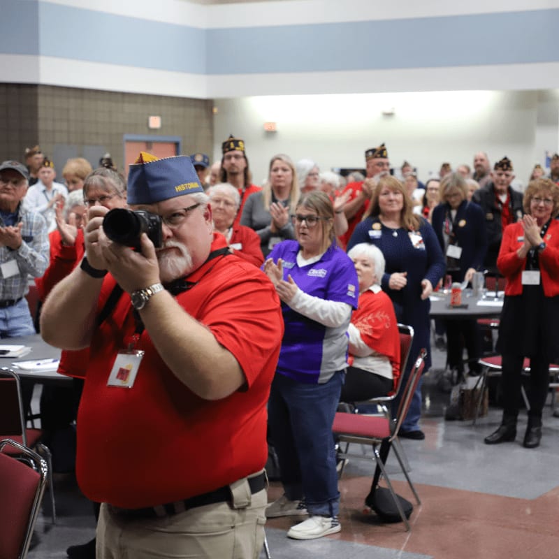 A man in a red shirt and blue hat with a camera in the foreground is surrounded by people in the background, many wearing hats and some clapping.