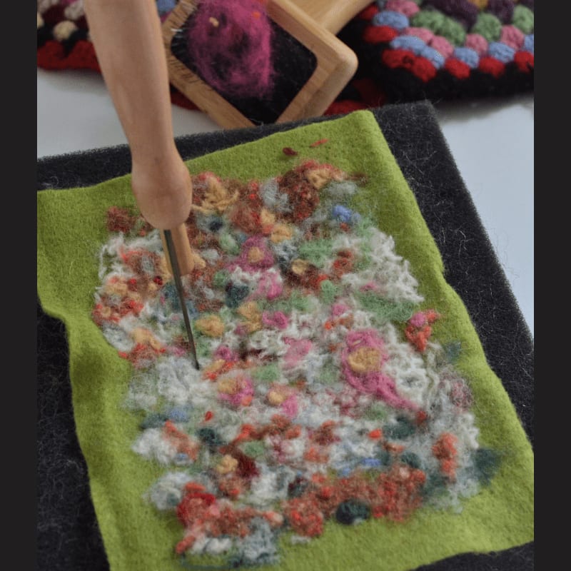 A needle tool is used to felt colorful fibers onto a green fabric in the foreground, while a wooden carder with pink wool and a crocheted blanket are visible in the background.