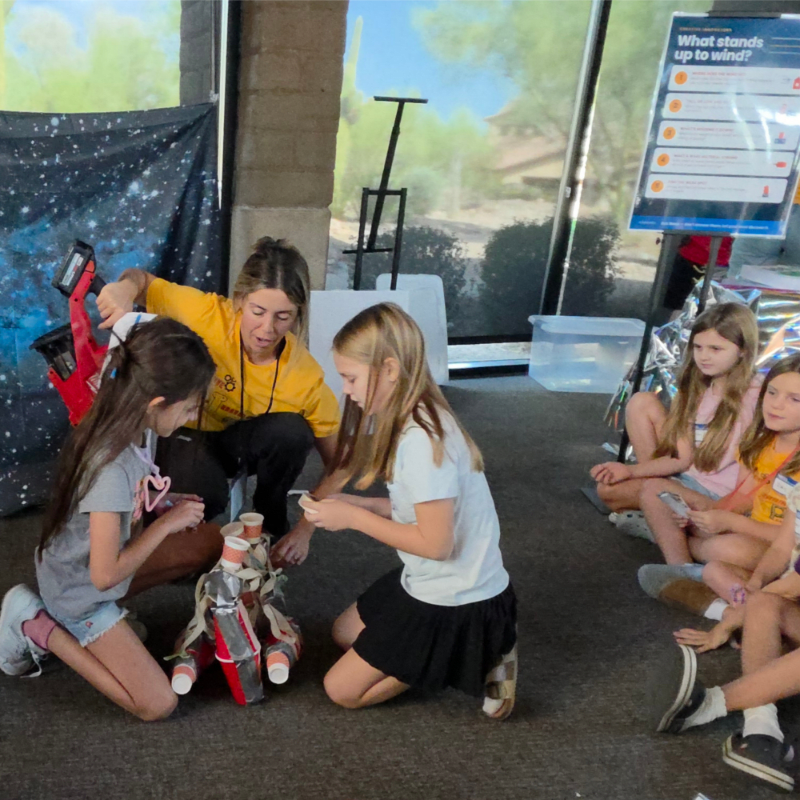 A woman in a yellow shirt guides young girls in a craft project in the foreground, while a display board and outdoor scenery are visible in the background.