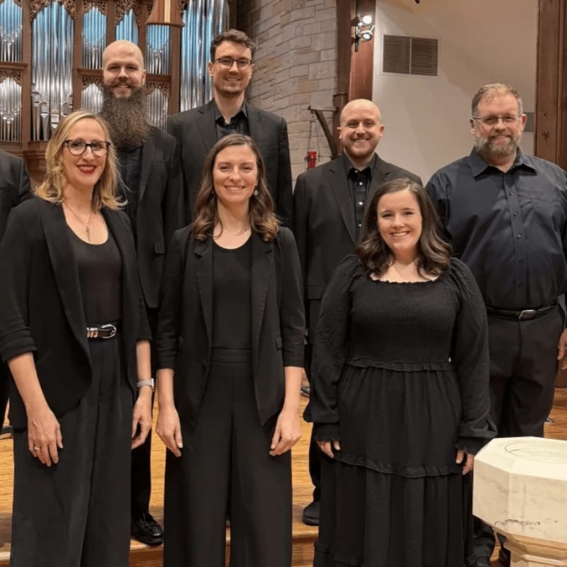 A choir of seven people in black attire stands in front of a pipe organ in a church.