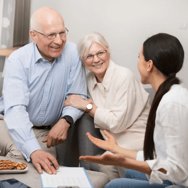 An elderly couple smiles warmly at a younger woman who gestures while speaking, with a plate of snacks on a coffee table in the foreground and a softly lit room in the background.