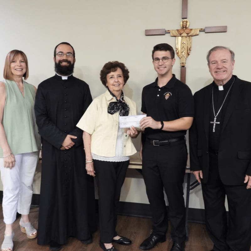 Five people stand in front of a wall with a large cross, with a woman in the center holding a check.