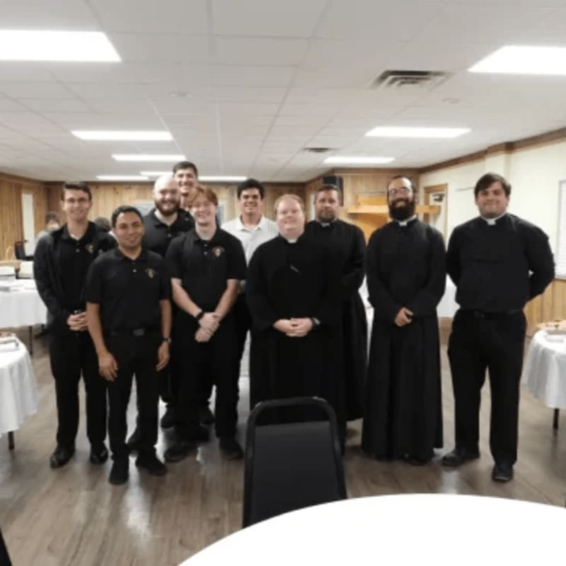 A group of men in clerical attire stand in a room with tables set up in the background.