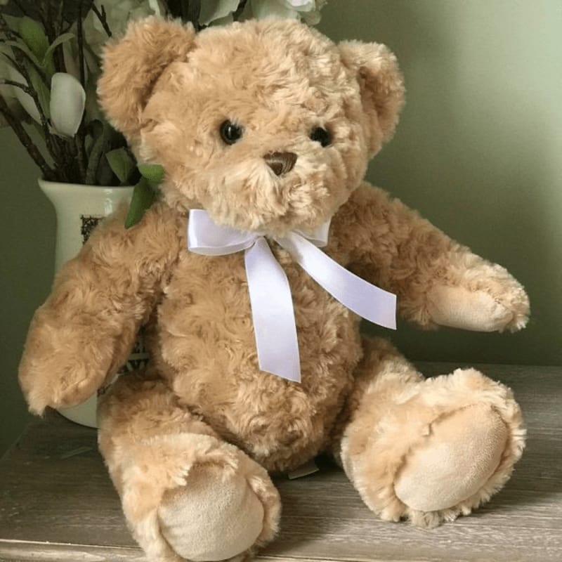 A fluffy tan teddy bear with a white ribbon bow sits in the foreground, with a white vase of greenery in the blurred background.